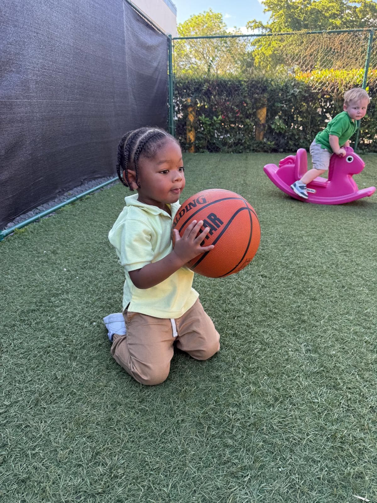 A child holding a basketball outdoors, with another child playing in the background