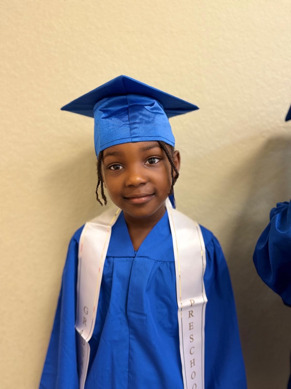 A child wearing a graduation cap and gown with a white “Preschool Graduate” sash